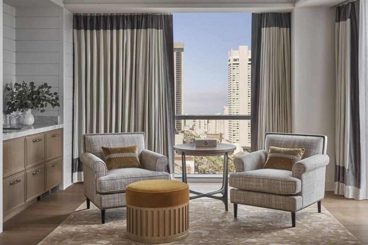 Ein modernes Wohnzimmer mit zwei Sesseln, einem kleinen runden Tisch und einem Hocker. Das Zimmer bietet große Fenster mit Vorhängen, einen Blick auf hohe Gebäude und eine Holzkommode.