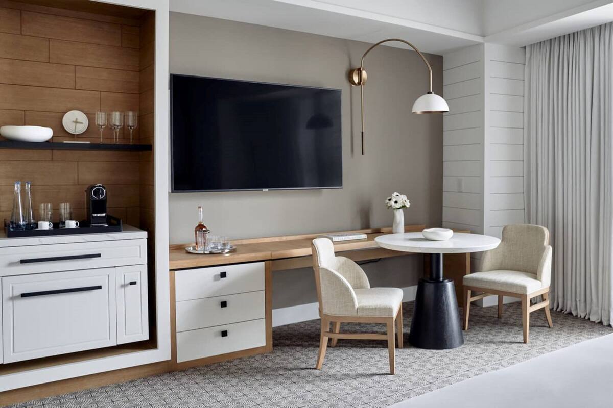 Ein modernes Hotelzimmer mit Flachbildfernseher, einem Holztisch mit Kaffeemaschine, zwei Stühlen, einem runden Tisch und einem großen Fenster mit Vorhängen.