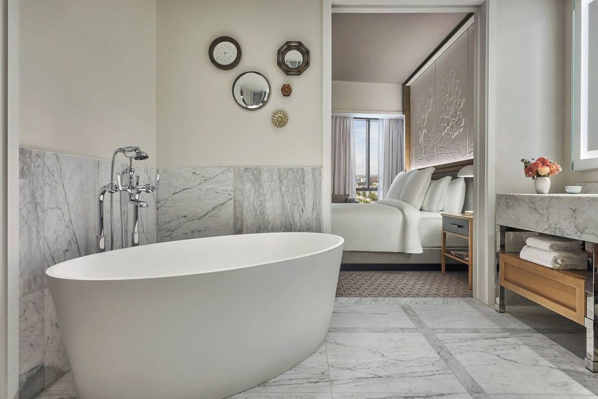Ein modernes Badezimmer mit freistehender Badewanne, Marmorwänden und Blick auf ein Schlafzimmer mit Bett und Fenster.