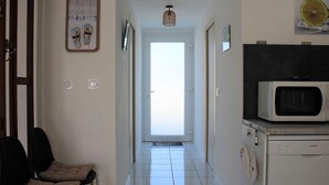 A hallway with a white door at the end, a microwave on a counter, and two chairs on the left side.