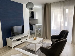 A modern living room with a flat-screen TV, a white coffee table with a decorative bowl, a dark armchair, and a dining area with a white table and chairs.