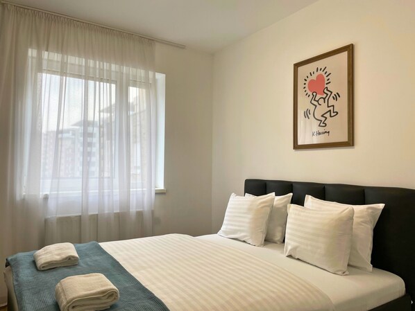 A hotel room with a bed, white curtains, a framed picture on the wall, and towels on the bed.