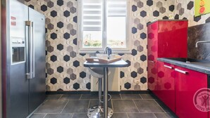 A kitchen with a hexagonal patterned wall, a stainless steel refrigerator, a red cabinet, and a window with blinds. - Single-level house with terrace, enclosed garden and pets allowed (Anneville-en-Saire)