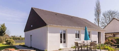 Uma casa térrea com telhado de telhas, um pátio com mesa e cadeiras, e um muro de pedra em primeiro plano.