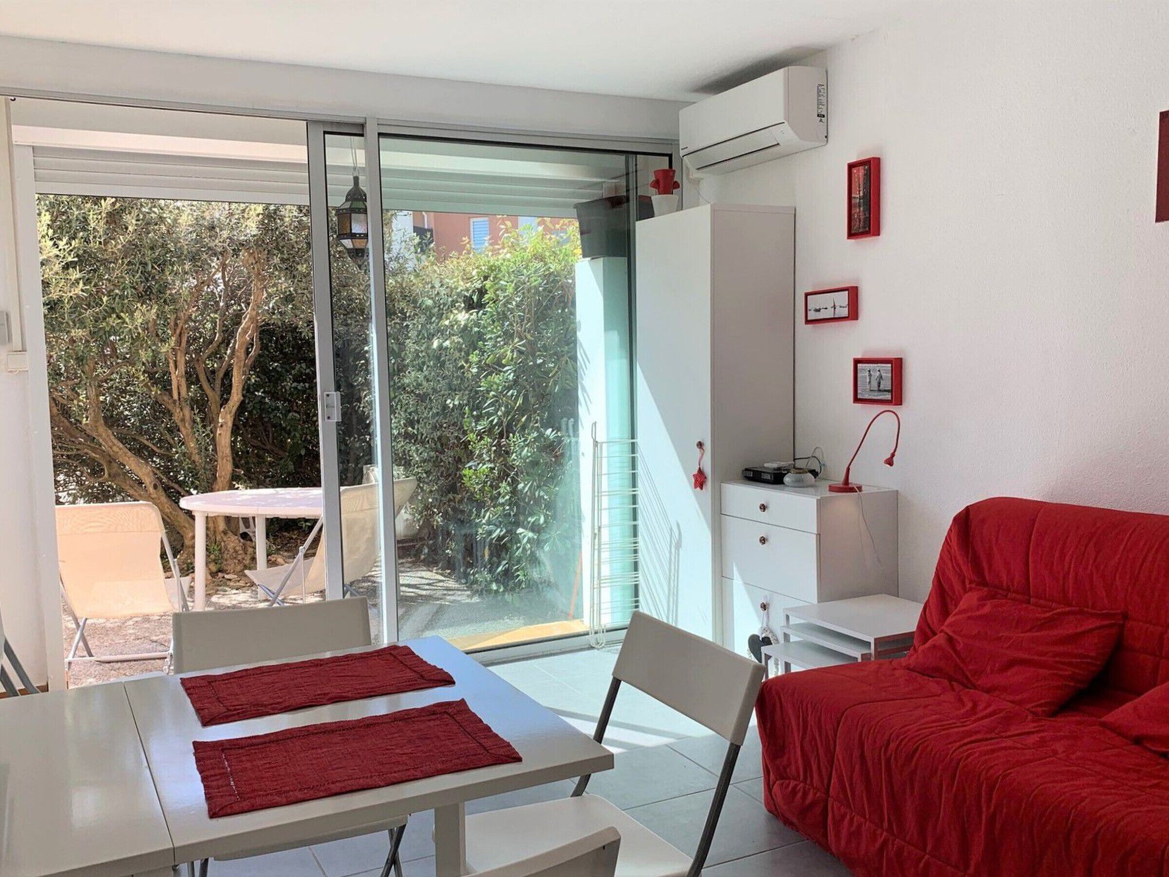 A modern room with a sliding glass door leading to a patio, a red sofa, a white table with red placemats, and a small white desk with a lamp.