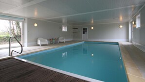 En indendørs swimmingpool med træterrasse, hvide vægge og en dør, der fører udendørs.