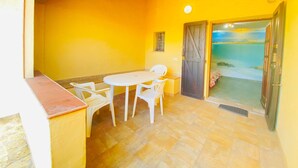 Una habitación con paredes amarillas, suelo de baldosas, una mesa blanca con sillas, una puerta que da a un balcón y una ventana pequeña.