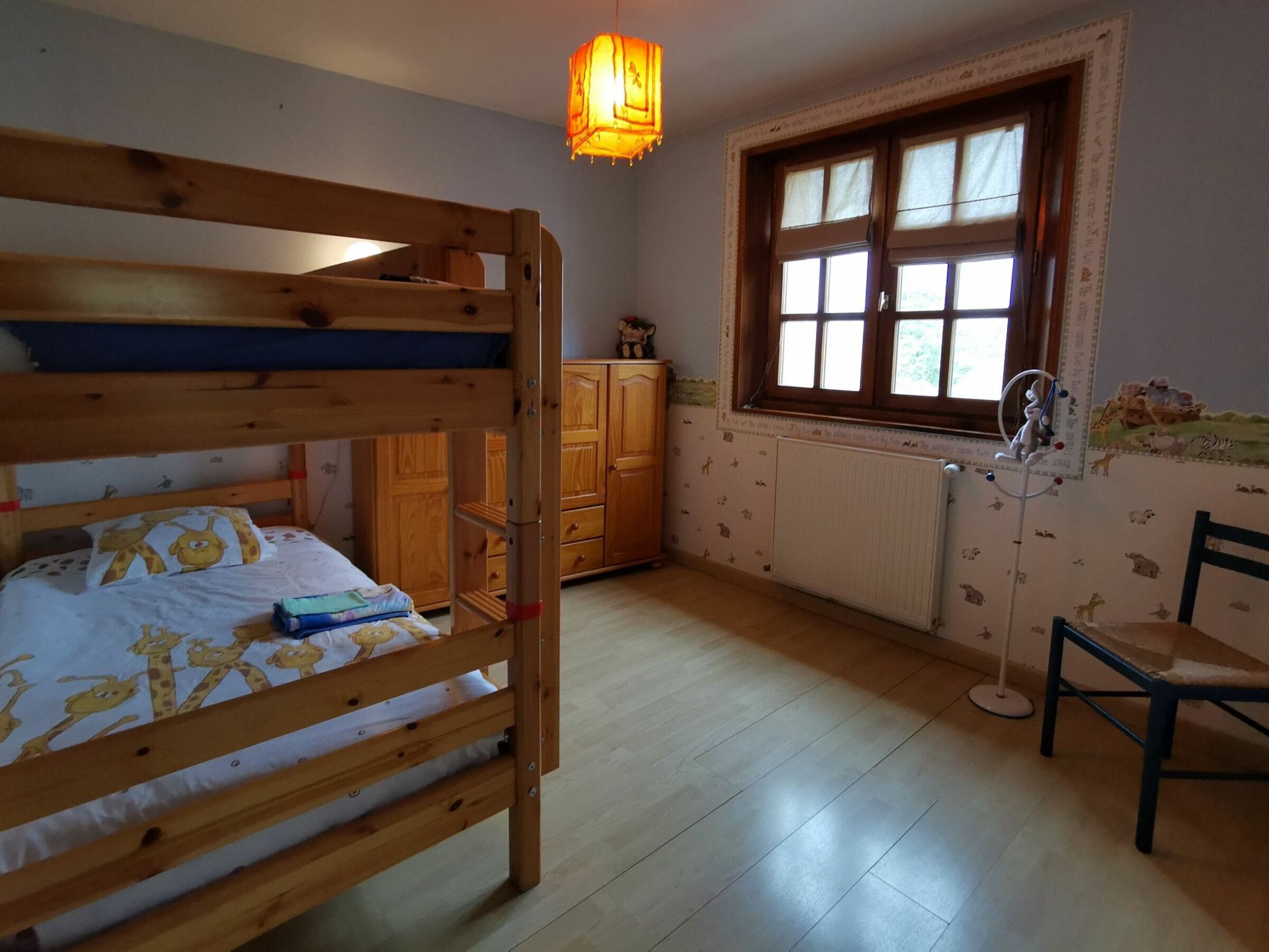 Une chambre avec un lit superposé, des armoires en bois, un radiateur, une fenêtre avec des rideaux et une chaise.