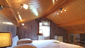 Une chambre confortable située sous les toits, avec un plafond en bois, un grand lit recouvert d’une literie à rayures, une table de chevet avec une lampe et une fenêtre agrémentée de rideaux.