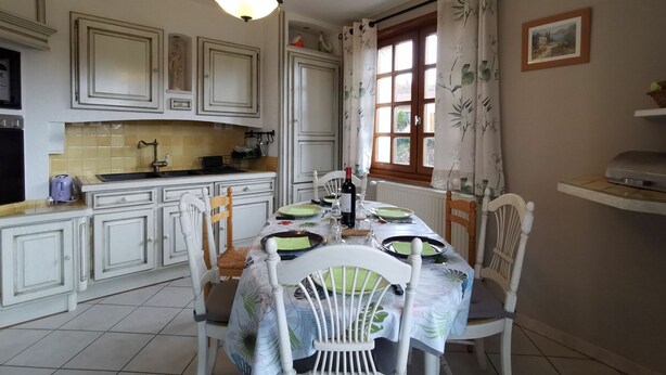 Un coin repas avec une table dressée pour quatre personnes, entourée de chaises blanches. Un lustre est suspendu au plafond, et une fenêtre avec des rideaux est visible.
