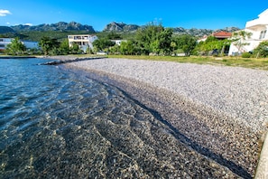 A pebbled beach with clear water, surrounded by buildings and mountains in the background. - Apartment with a garden near the beach (Seline)