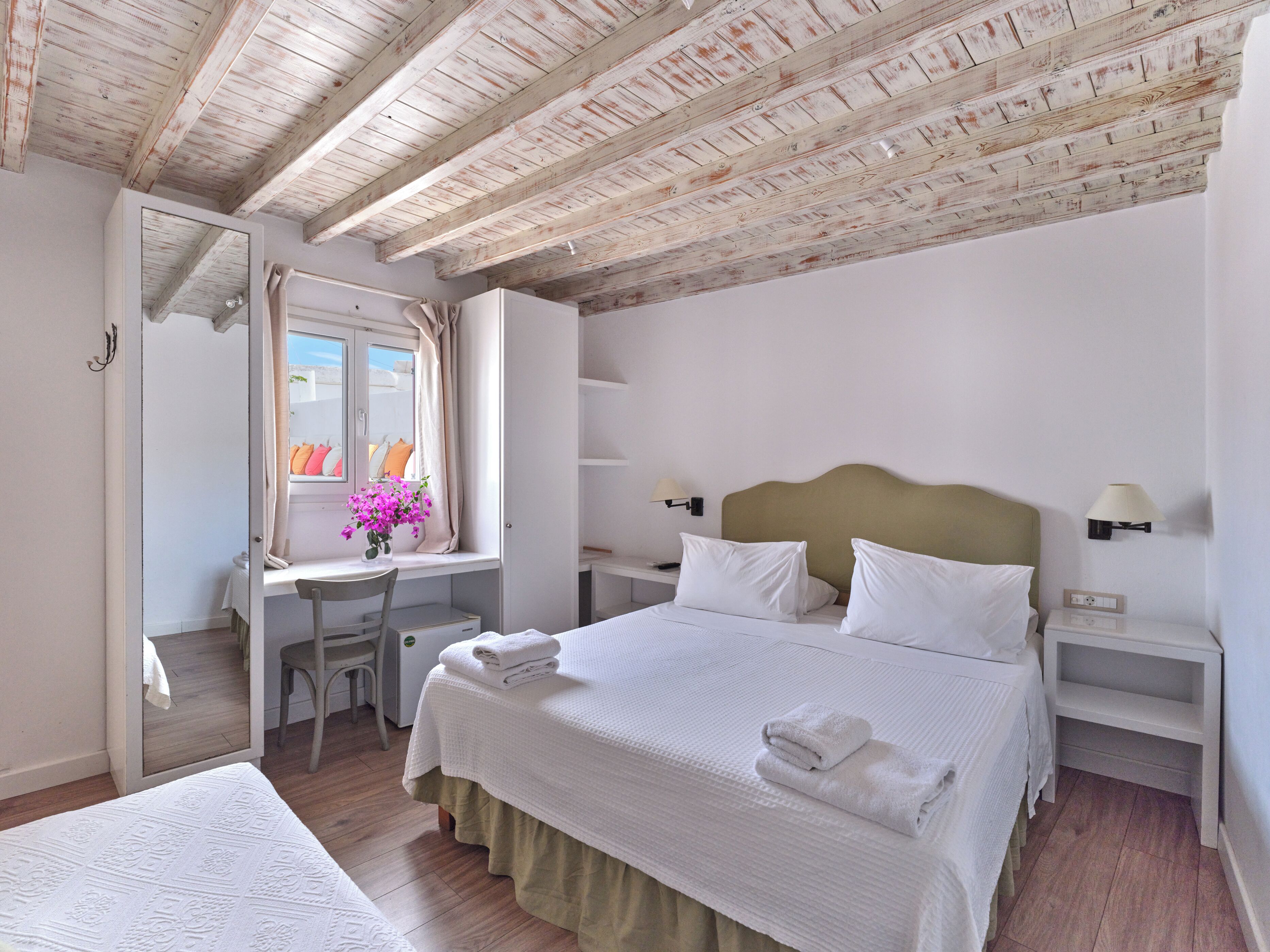 Un dormitorio con cama, escritorio, silla, ventana con cortinas y mesita de noche con lámpara.