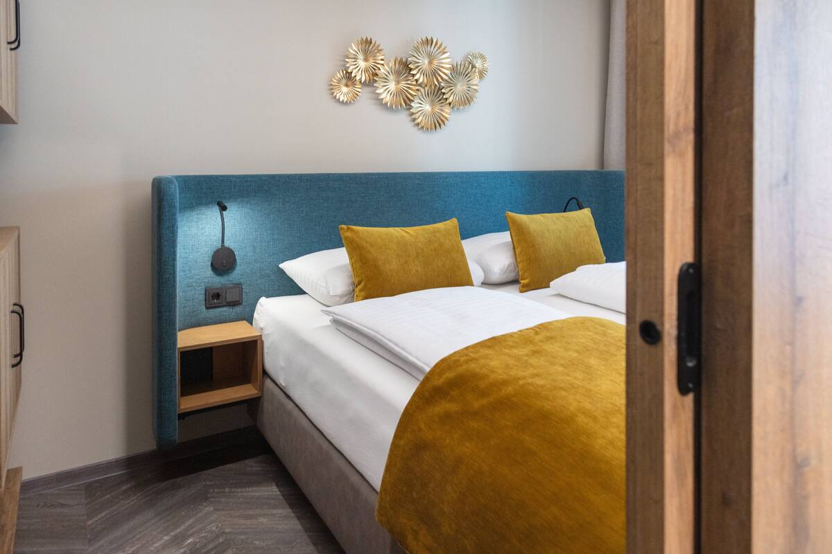 Una cama bien hecha con un cabecero azul, dos almohadas color mostaza y una mesita de noche de madera con una lámpara.