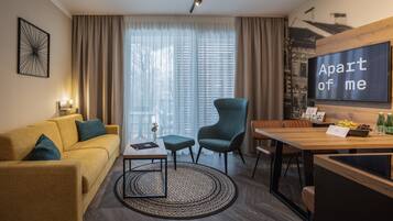 A modern hotel room with a sofa, a chair, a dining table, a TV, and a large window with curtains.