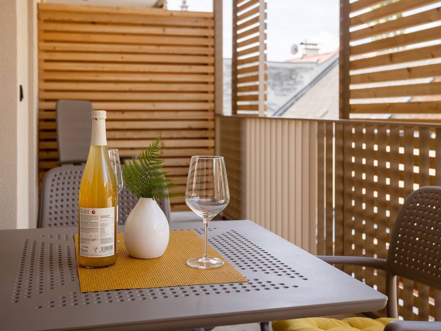 A table with a bottle of wine, a glass, and a small white vase with a plant on a balcony with wooden slats.