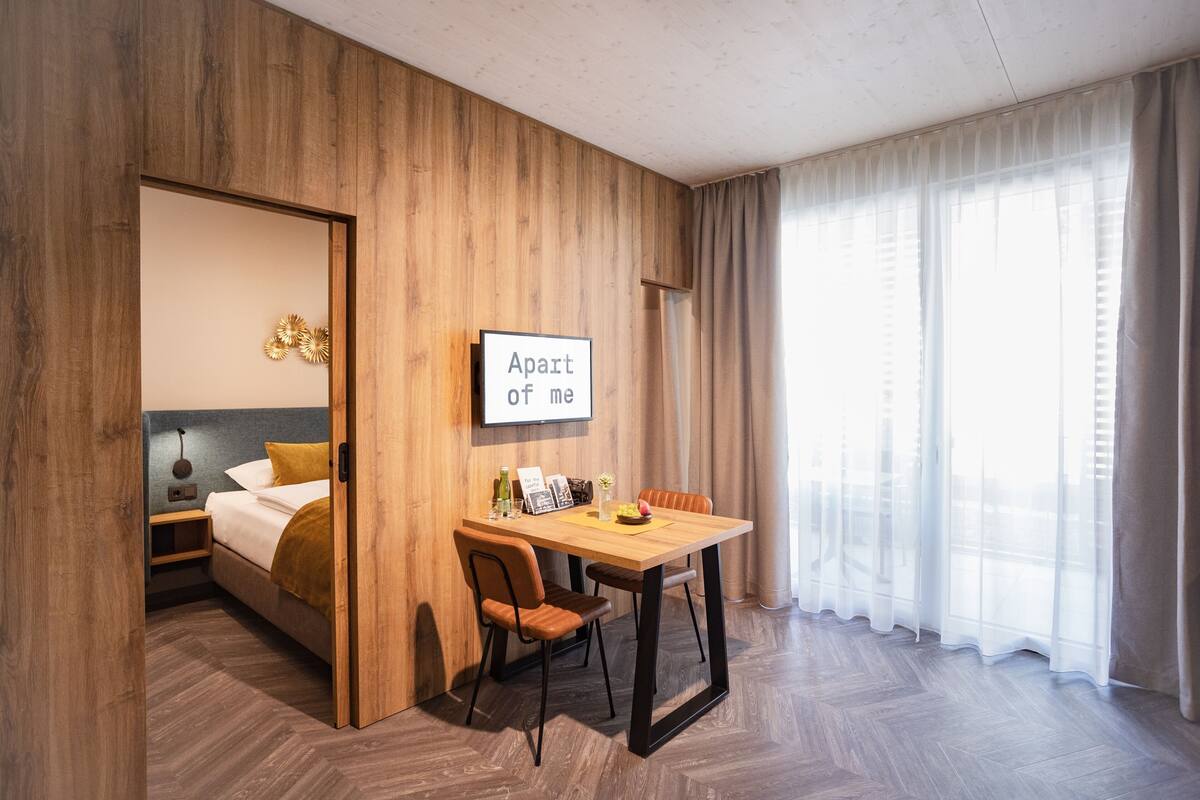 Habitación de hotel con pared de madera, una cama con almohada amarilla, una mesa de comedor con sillas y un ventanal con cortinas traslúcidas.