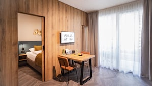 A hotel room with a wooden wall, a bed with a yellow pillow, a dining table with chairs, and a large window with sheer curtains.