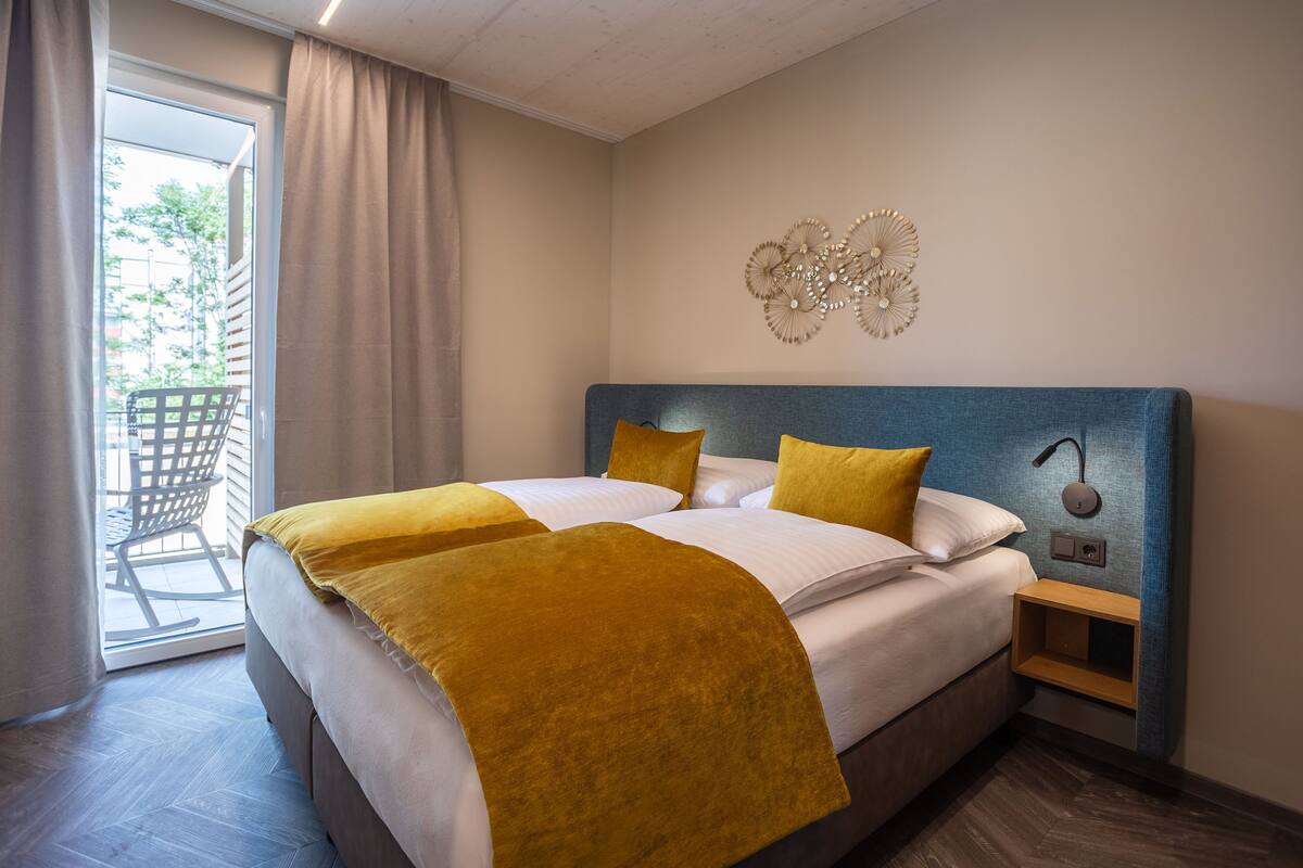 Habitación de hotel con una cama grande, una mesita de noche, un reloj de pared y vistas al exterior a través de una puerta corredera.