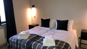 A neatly made bed with white linens, black and grey checkered throw, and two folded towels on the foot of the bed. There are two bedside tables, one on each side of the bed, with a small lamp on each. The room has a window with dark curtains, and a radiator is visible on the wall. - Hellnar Ocean View Villa (Hellnar)