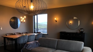 A modern living room with a sofa, dining table, chairs, and a large window offering a view of the outdoors. - Hellnar Ocean View Villa (Hellnar)
