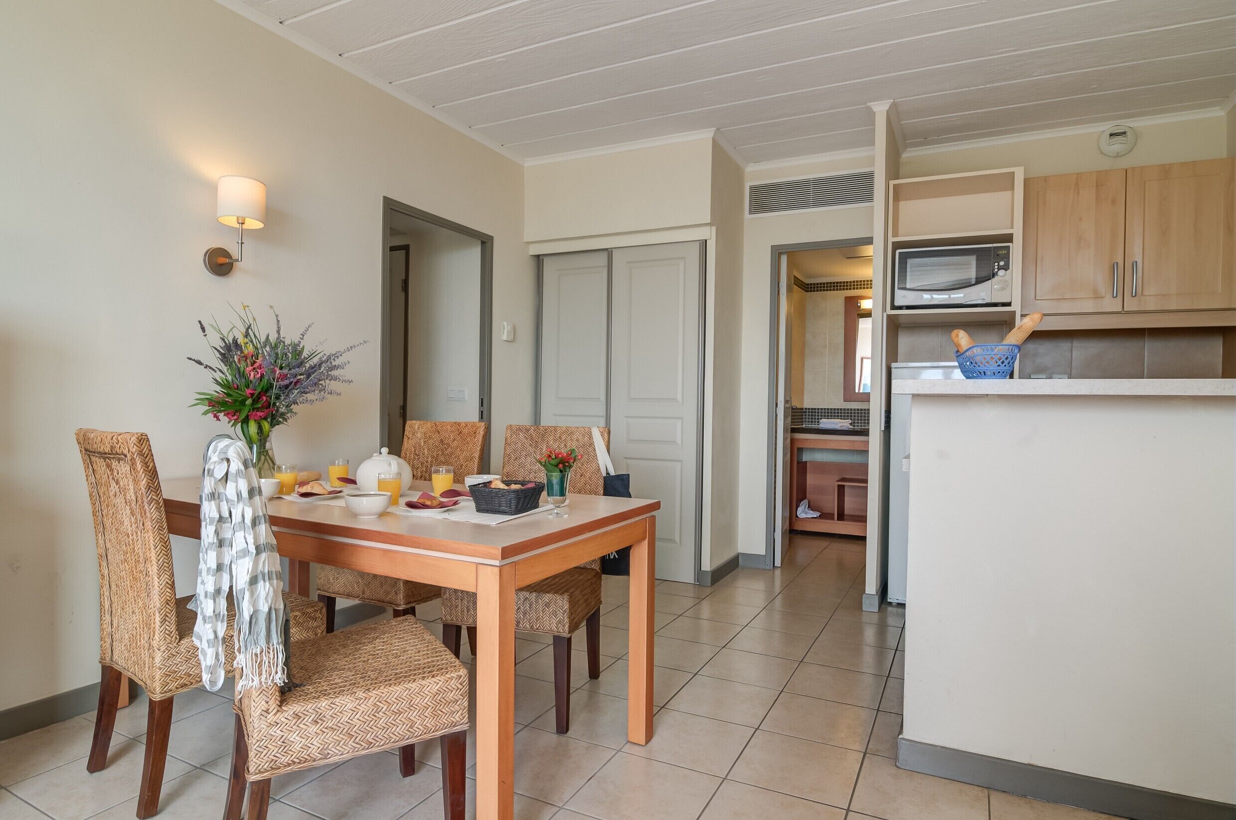 A dining area with a wooden table, chairs, and a vase of flowers. There is a microwave and a basket on the counter.