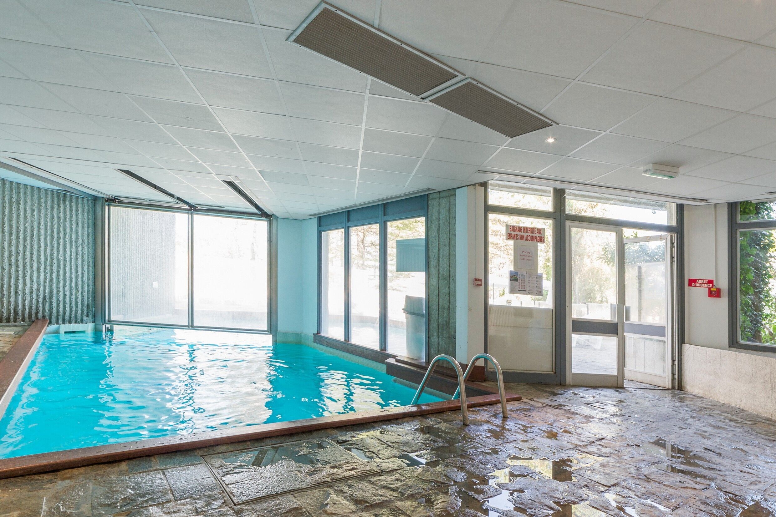 An indoor swimming pool with clear glass walls, a tiled floor, and a metal ladder.