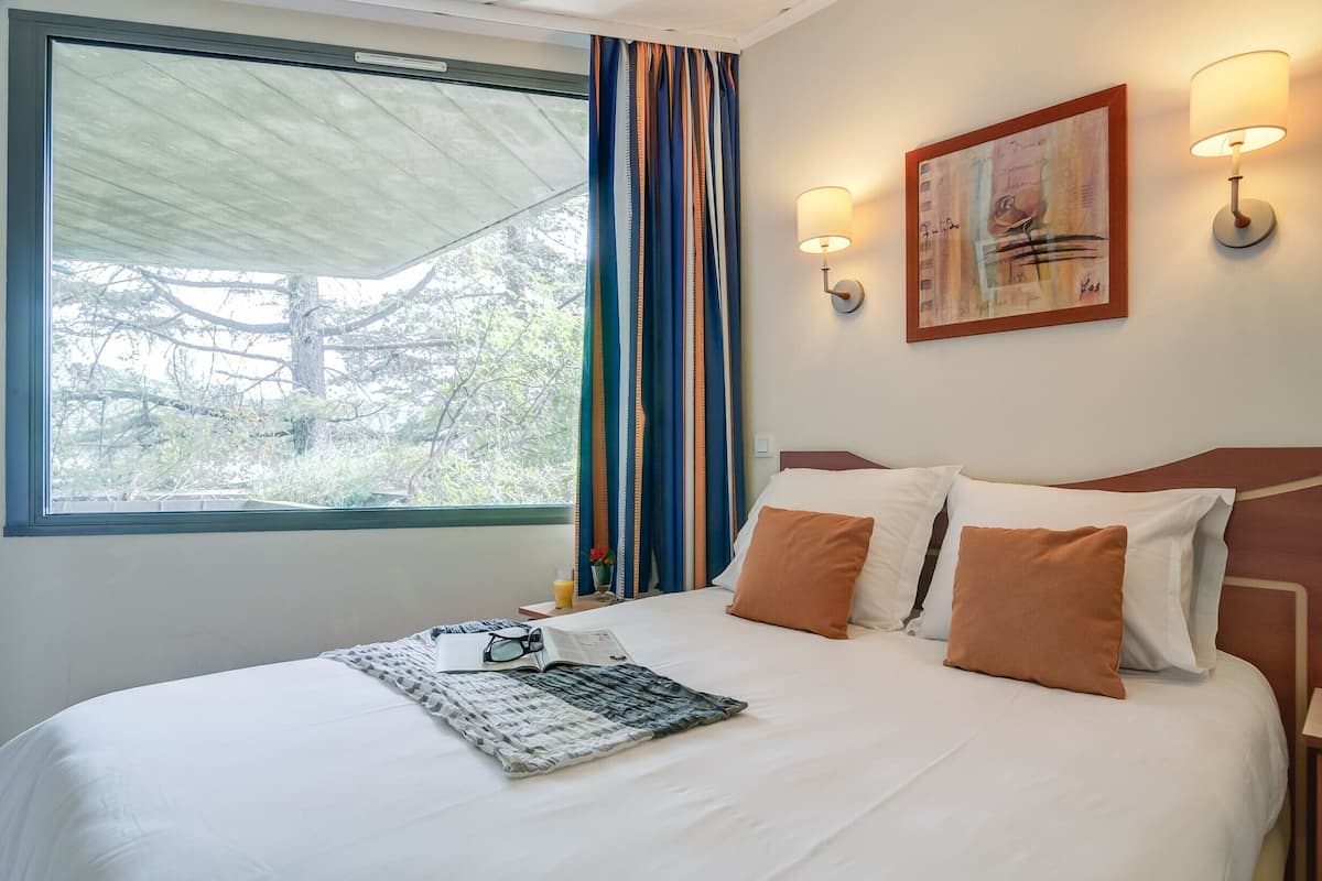 A neatly made bed with a magazine, glasses, and a bottle of water. There is a window with a view of trees and a curtain.
