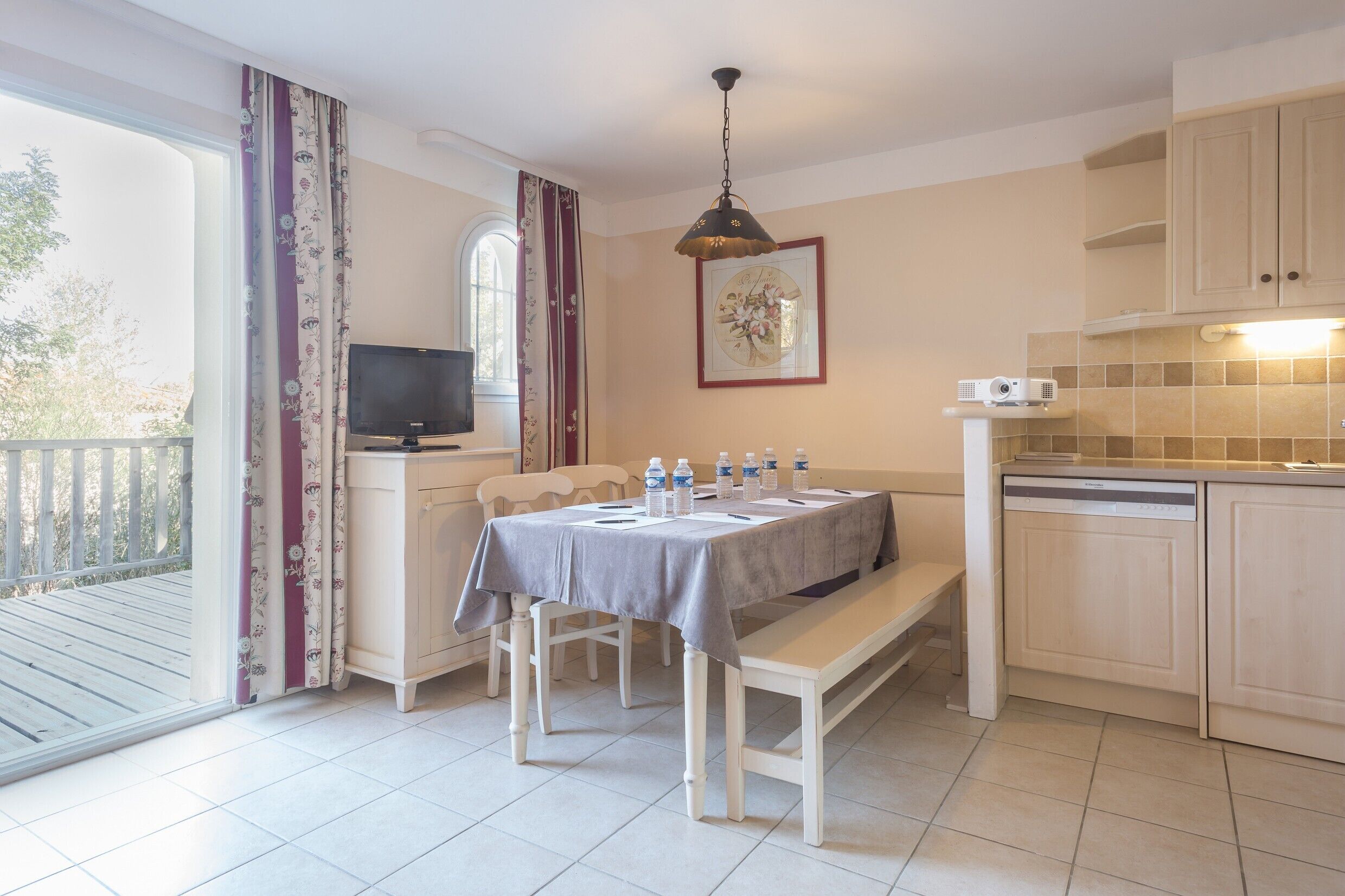 Un coin repas avec une table dressée pour deux personnes, un banc, un téléviseur et un espace cuisine équipé de placards et d’un lave-vaisselle.