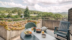 A balcony with a table set for breakfast, including fruits, pastries, and beverages, overlooking a lush landscape with hills and trees. - SOWELL HOTELS Ardèche (Grospierres)