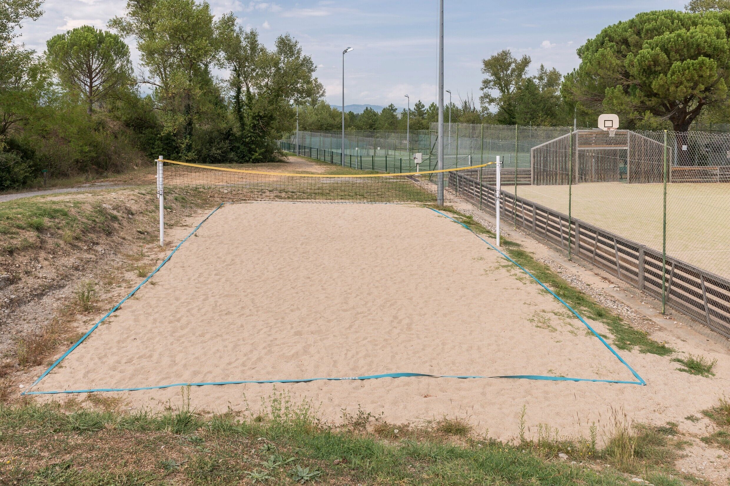 Un terrain de volleyball de sable avec un filet, entouré d’une clôture et d’arbres.