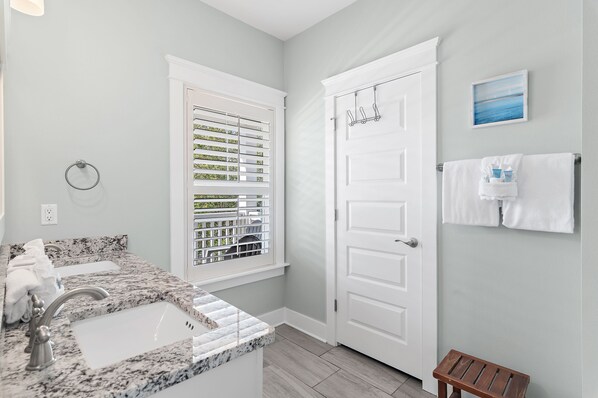 A bathroom with a granite countertop sink, a window with blinds, a white door, and a small wooden stool.