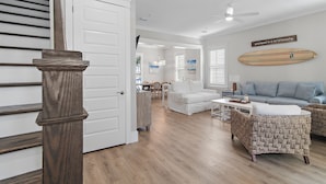 A spacious living area with a white sofa, wicker chairs, a dining table, and a surfboard decoration on the wall.