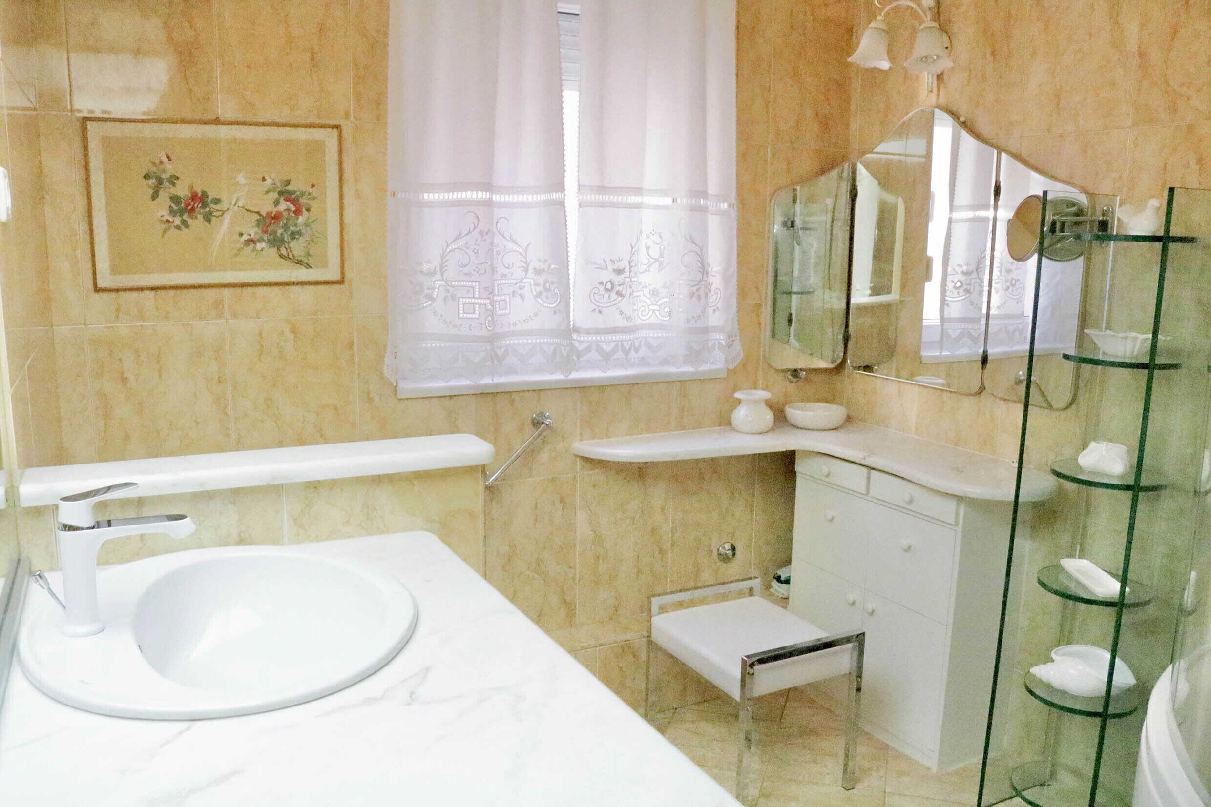 A bathroom with a white sink, a small step stool, a vanity with a mirror, and a glass shower enclosure.