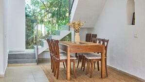 A dining area with a wooden table and chairs, a vase with dried flowers, and a view of greenery through large windows.