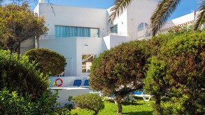 A modern white building with a pool area, surrounded by well-maintained greenery and palm trees.