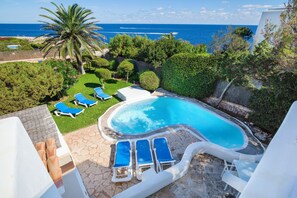 A swimming pool with lounge chairs and a palm tree, surrounded by greenery and a view of the sea.