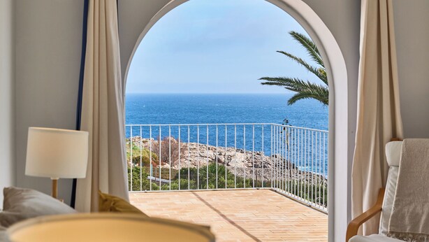 A room with a view of the sea through an arched window, a balcony with a railing, and a chair with a towel.
