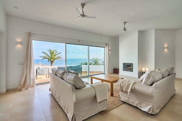 A spacious living room with a large sliding glass door leading to a balcony with a view of the sea. There are two beige sofas, a wooden coffee table, and a ceiling fan.
