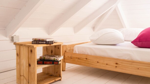 Een houten bed in een kamer met schuine plafonds en een klein houten nachtkastje met boeken.