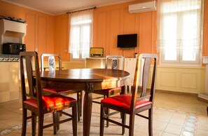 A dining room with a wooden table and six chairs with red cushions. There is a television mounted on the wall and a microwave on a shelf.