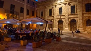 An outdoor café with tables and umbrellas, surrounded by stone buildings with arched doorways and windows. - Best Located in Alghero old Town Steps to sea Attic Emerald (Alghero)