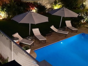 Uma área à beira da piscina com espreguiçadeiras e guarda-sóis, uma mesinha e uma piscina.