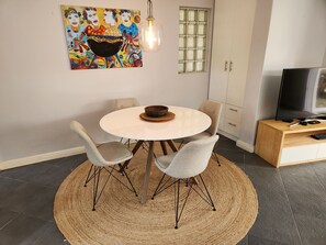 Un coin repas comprenant une table ronde, quatre chaises, un tapis tissé, un tableau, une fenêtre avec des stores, un téléviseur posé sur un meuble en bois et un buffet.