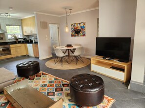 A modern kitchen with wooden cabinets, a dining area with a round table and chairs, a television on a wooden stand, and a colorful rug. - Charming Fully Fitted Double Storey Cottage Near Constantia Village (Cape Town)