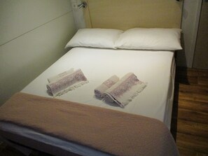 A single bed with white bedding, two rolled towels, and a brown blanket.