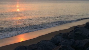 Ein Strand mit Sonnenuntergang über dem Wasser, felsigem Ufer und entfernten Bergen.