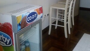 Quarto compacto com uma pequena geladeira, uma cadeira branca e uma porta de madeira.