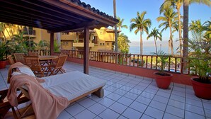 A terrace with a lounge area, including a bed and chairs, overlooking a beach with palm trees. - Villa Facing the Beach in a Large Terrace 7 Pools, Tennis Courts, Security (Puerto Vallarta)