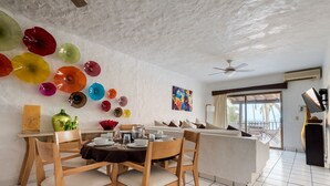 A dining area with wooden chairs and a table set for four, a white sofa, a ceiling fan, and a large window with a view of palm trees. - Villa Facing the Beach in a Large Terrace 7 Pools, Tennis Courts, Security (Puerto Vallarta)