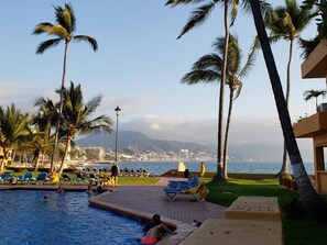 A pool area with lounge chairs, palm trees, and a view of the city and mountains in the background.